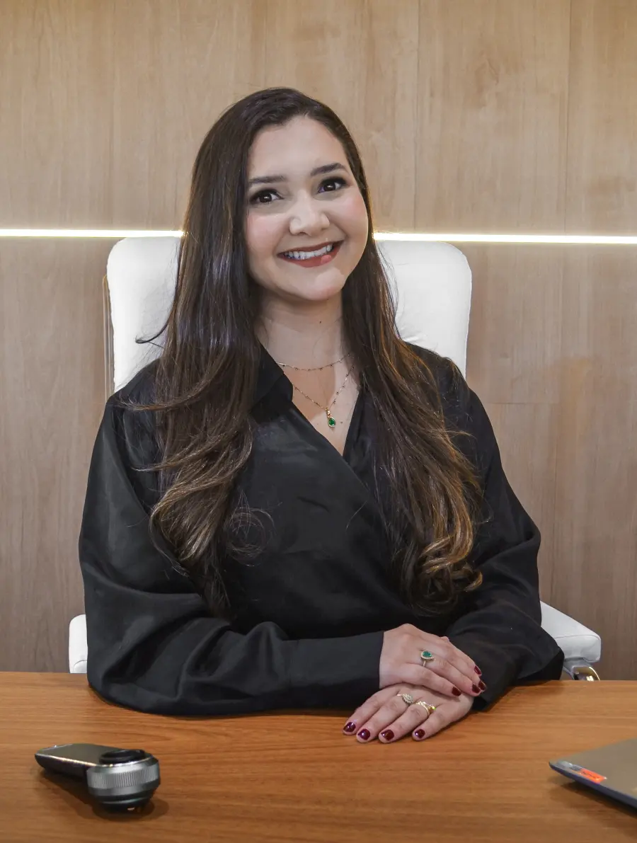 Dra. Ester Santana sorrindo, sentada à mesa do consultório
